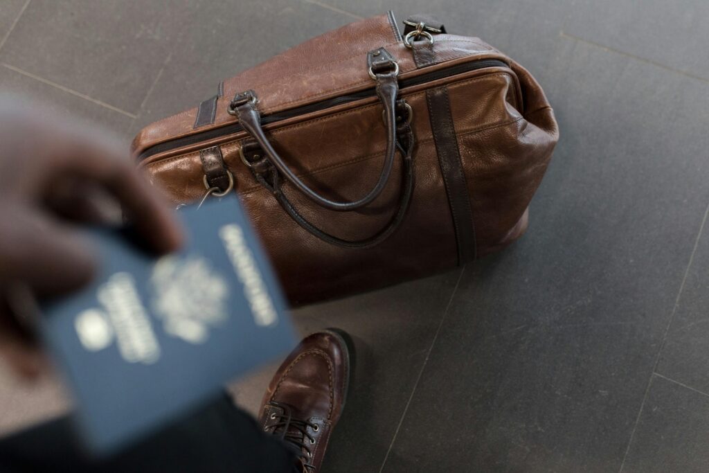 A passport is held above a brown leather travel bag on the floor