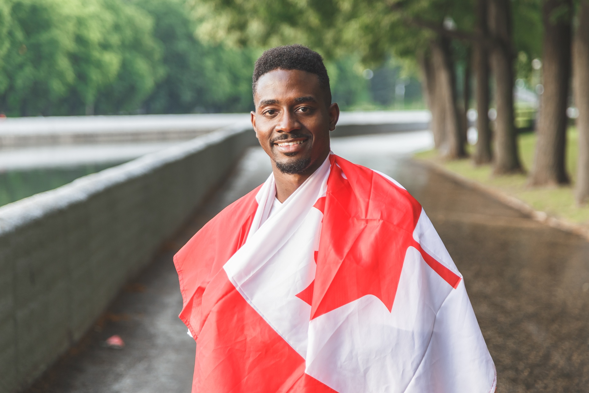 Handsome man with Canadian flag on his shoulders smiling and looking at camera, standing outdoors